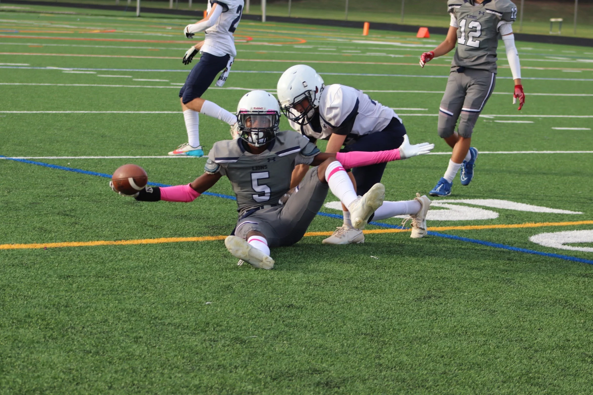 A football player in a gray uniform with the number 5 is sitting on the ground, holding a football, while a teammate in a white uniform is reaching toward him. Two other players in gray and white uniforms are nearby on a green football field.