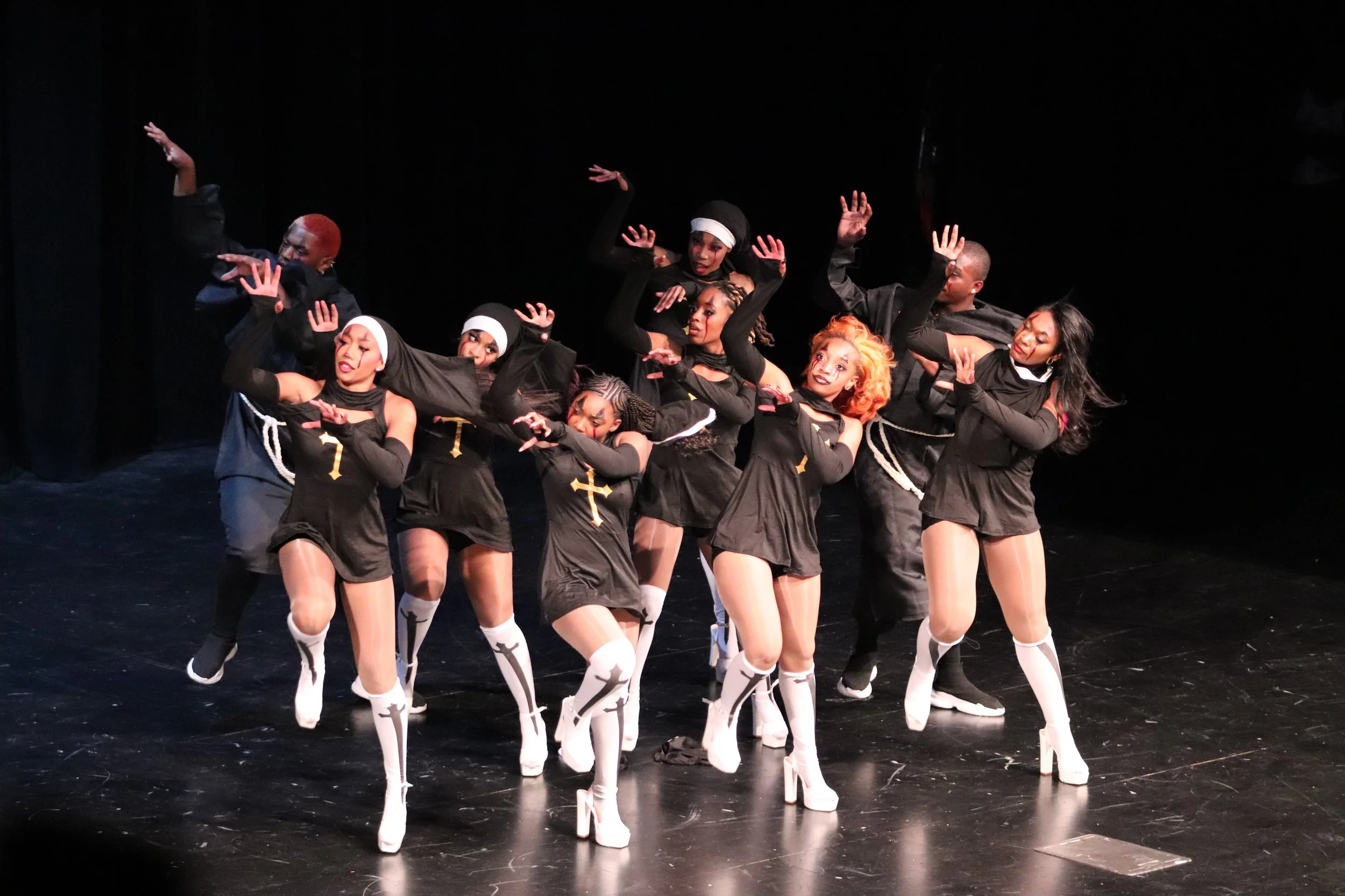 Group of performers on stage dressed in black, with some wearing nun costumes, sneakers, and stockings with crosses, dancing with synchronized arm movements.
