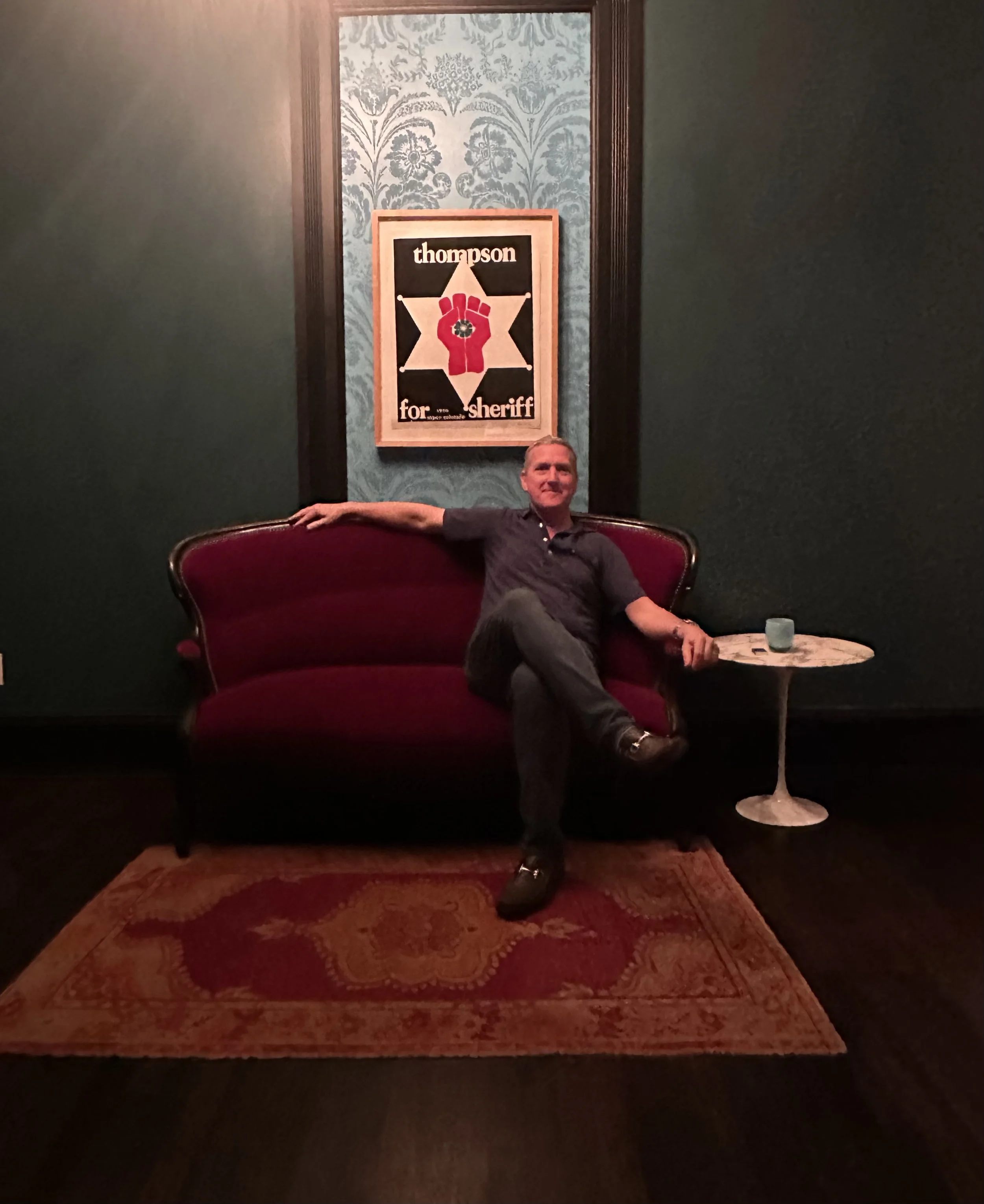 A man sitting on a red velvet sofa in a dimly lit room with vintage wallpaper, next to a small round table with a glass and a folded paper, and a framed sign that reads 'Thompson for Sheriff' with a sheriff's badge in the background.