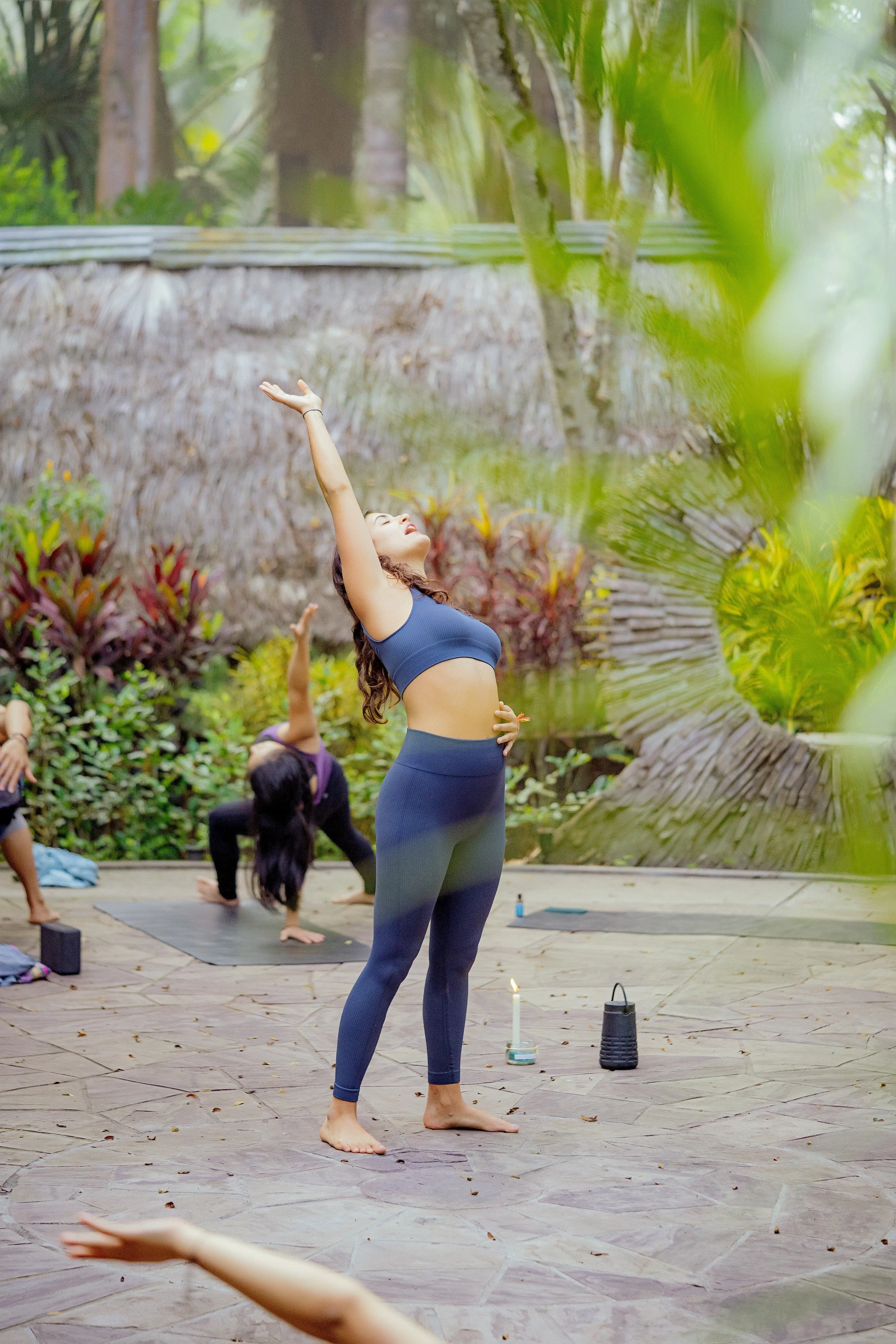 A woman doing yoga outdoors on a stone patio, with other people practicing yoga behind her. She is standing with one arm raised, eyes closed, and appears peaceful. The background includes lush green trees and plants.