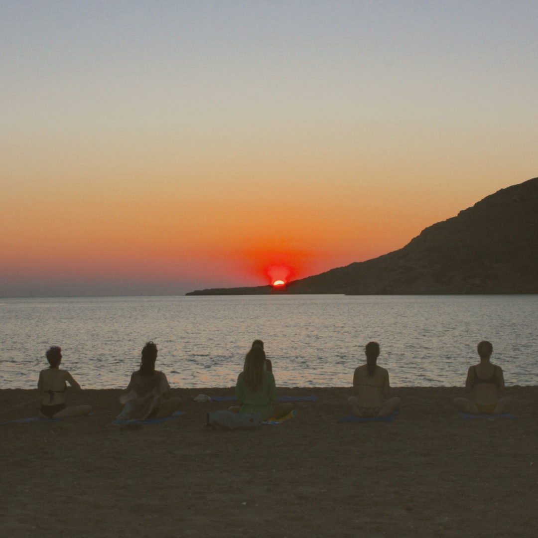 Five women practicing yoga on a beach during sunset.