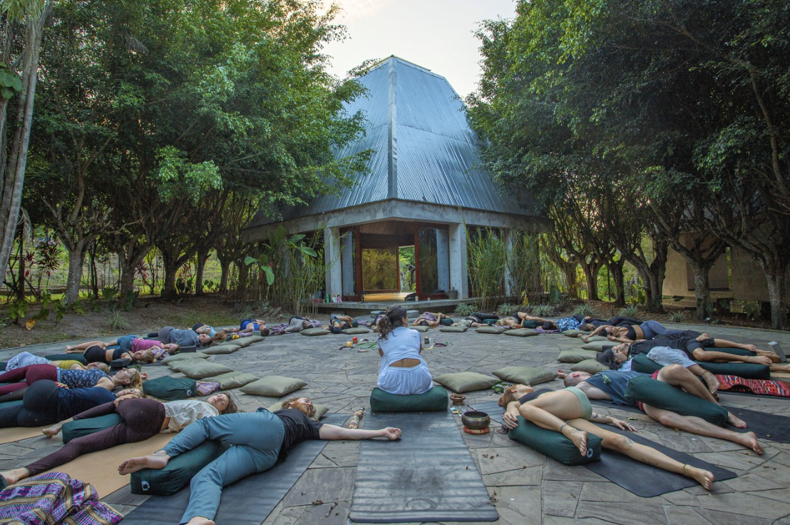 Group of people practicing yoga outdoors on mats, lying on their sides with some in a relaxation pose, under trees near a modern building with a metal roof.