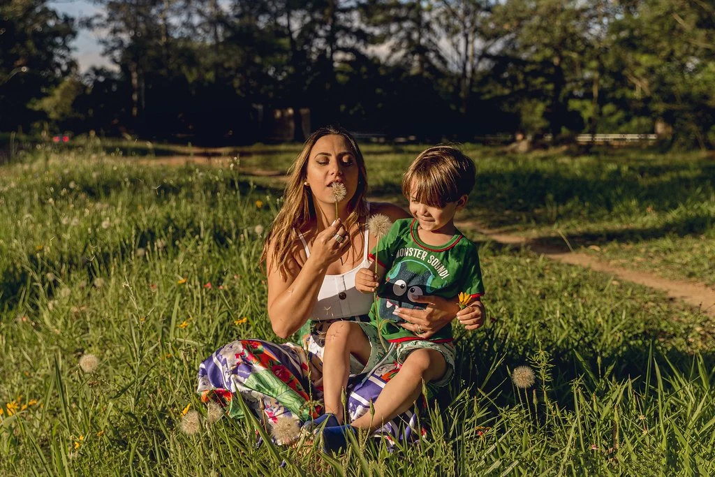 Mulher e criança sentados na grama, brincando e segurando dente-de-leão em um campo ao ar livre, com árvores ao fundo.