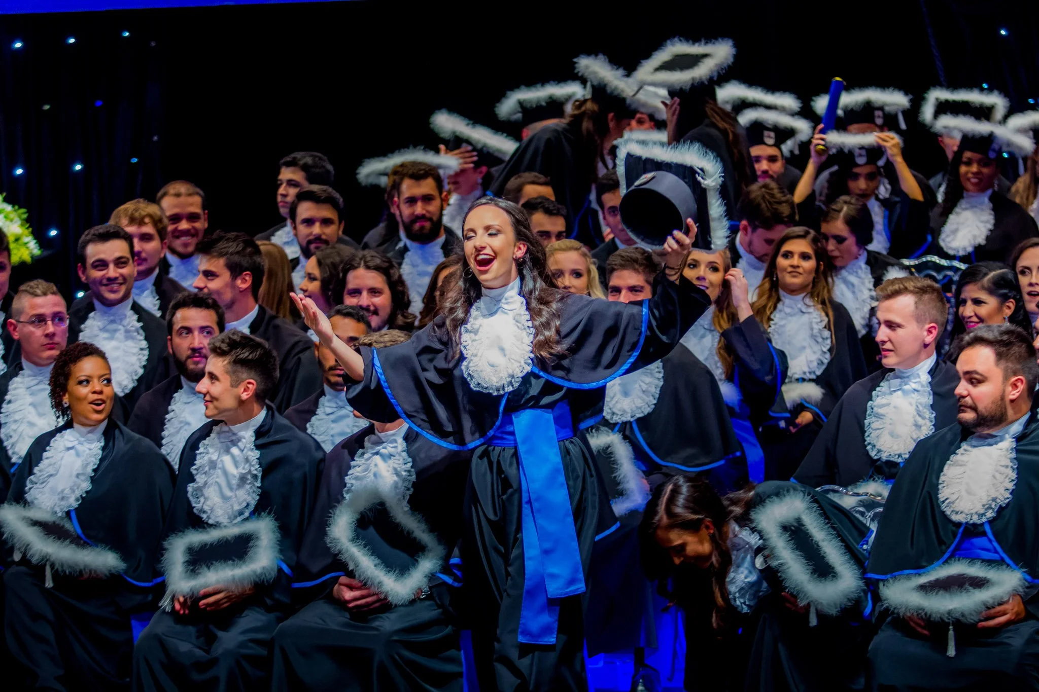 Graduação com pessoas vestindo beca e chapéu, uma mulher ao centro sorrindo, segurando o chapéu no ar, celebração em ambiente fechado.