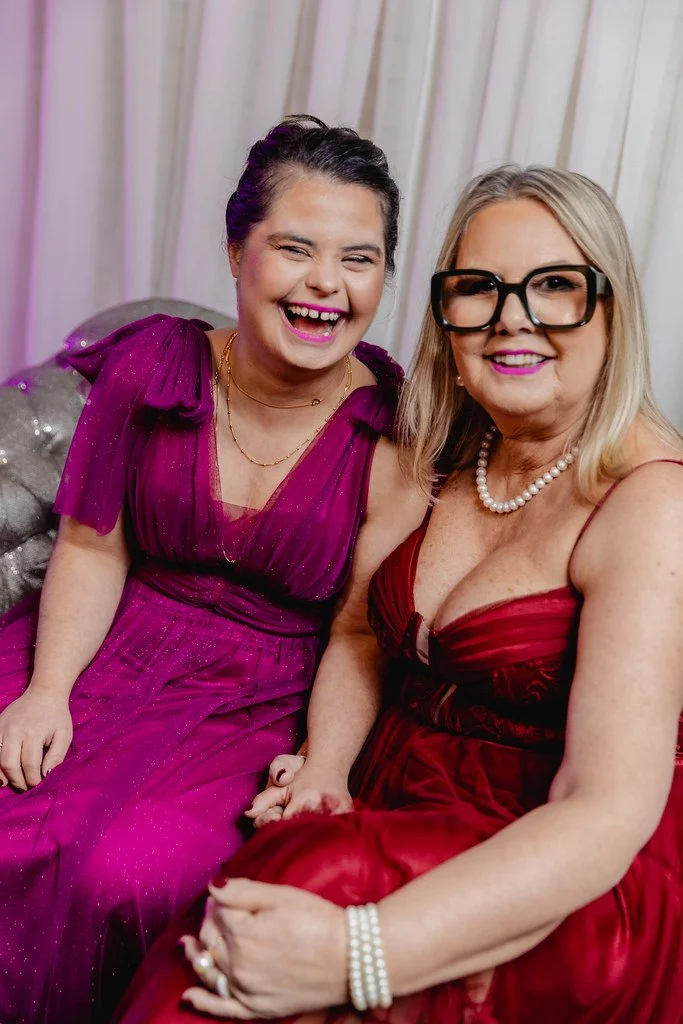 Duas mulheres sorrindo e vestidas com vestidos elegantes, uma de blusa roxa e outra de vestido vermelho, sentadas juntas em um ambiente com cortinas brancas ao fundo.