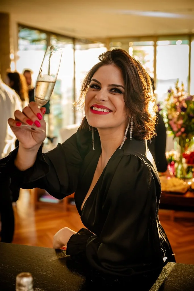 Mulher sorridente com cabelo castanho, usando vestido preto e brincos longos, segurando uma taça de champanhe em um ambiente bem iluminado, possivelmente uma festa ou celebração.