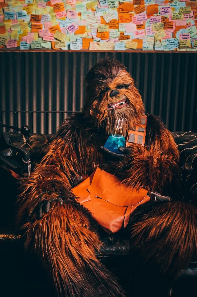 Pessoa vestida de Chewbacca segurando um copo com bebida azul, com mural de notas coloridas ao fundo.