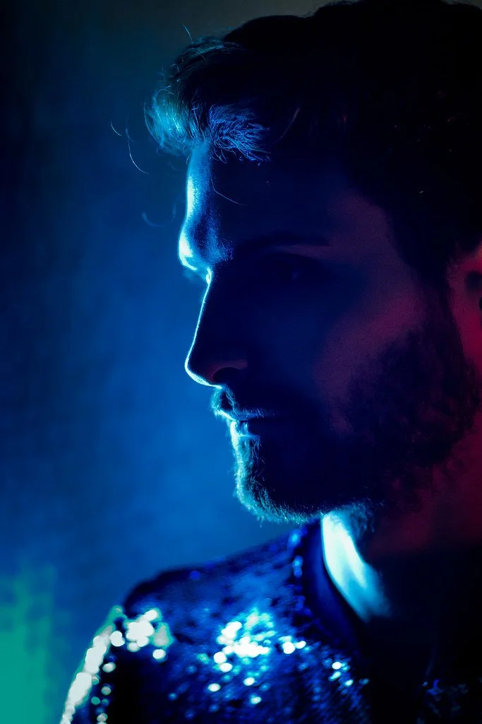 Retrato de um homem com iluminação azul e vermelha, com barba, usando jaqueta brilhante, em perfil.