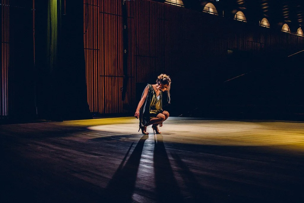 Mulher ajoelhada no palco, com iluminação dramática e fundo de painéis de madeira, em uma apresentação ou ensaio.