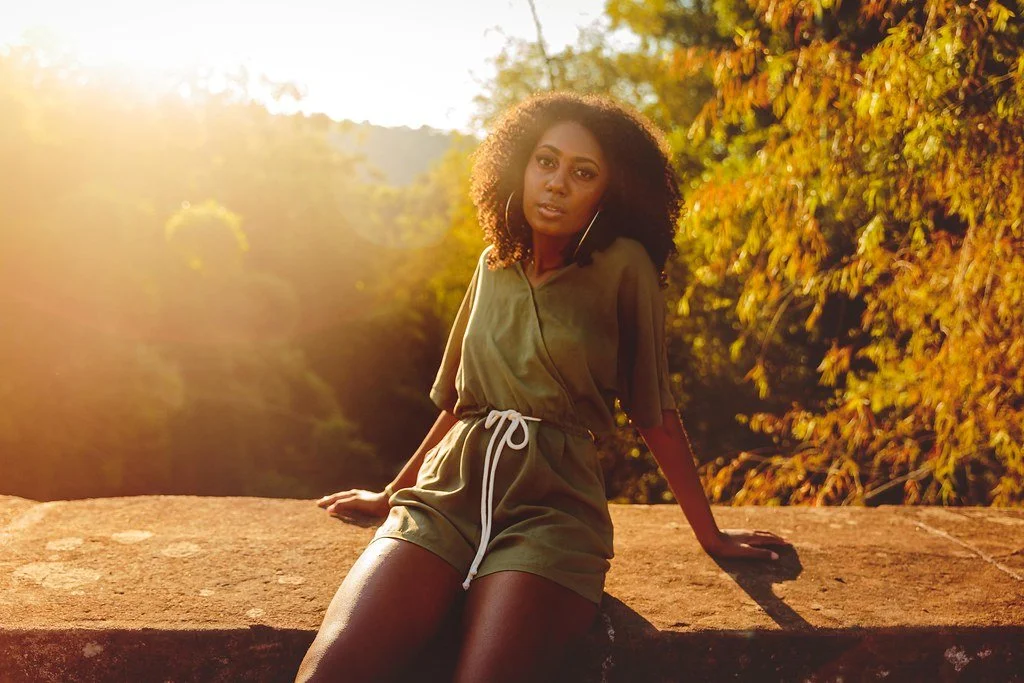 Mulher com cabelo cacheado de pé no muro durante o pôr do sol, com roupas curtas verdes e bege, ao ar livre.