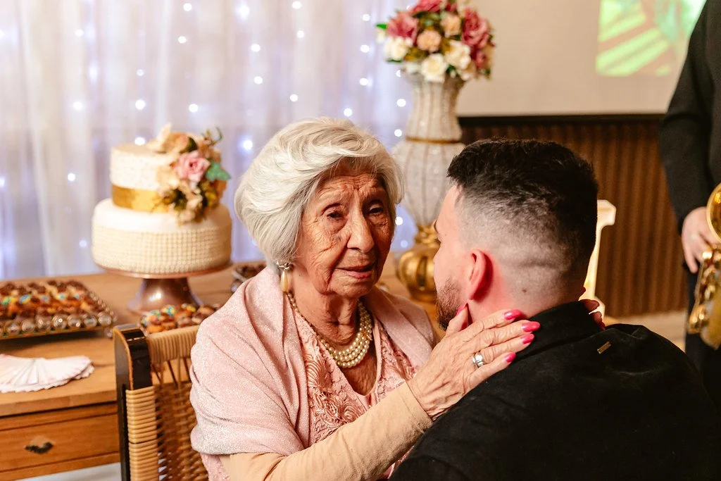 Uma senhora idosa com cabelo branco e vestida de rosa segurando o rosto de um jovem rapaz com cabelo curto e escuro, numa ocasião de celebração, ao fundo há uma mesa com bolo decorado com flores e uma decoração de iluminação com luzes brancas.