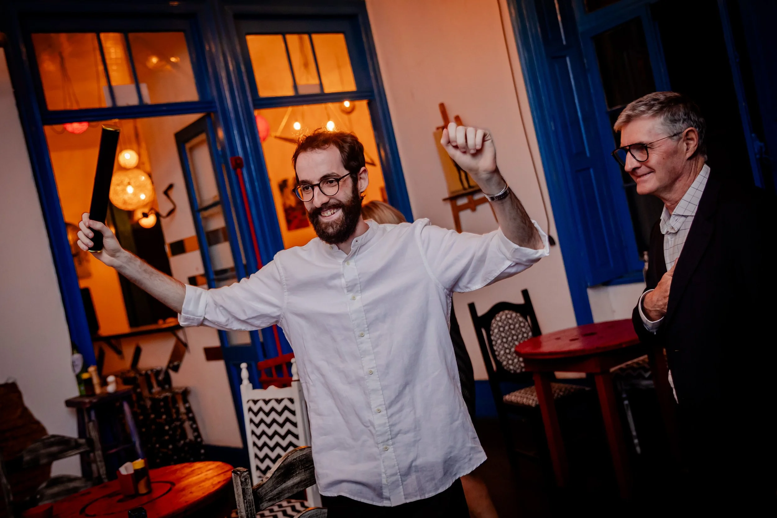 Dois homens celebrando em uma festa, um deles segurando um objeto preto na mão e dando um pulinho, ambos sorrindo, com luz quente e decoração colorida ao fundo.
