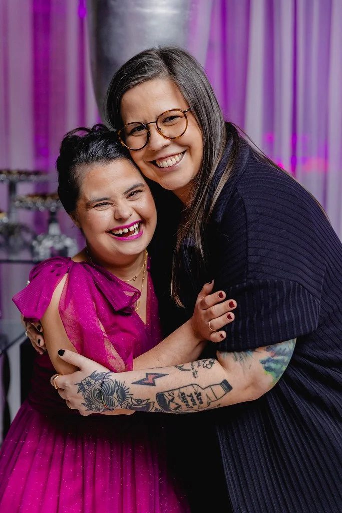 Duas mulheres sorrindo e se abraçando, uma com cabelo comprido e óculos, usando roupa preta, e outra com cabelo escuro, maquiagem marcante, vestido rosa com ombros de mangá bufante, e tatuagens visíveis no braço, em um ambiente com iluminação roxa.