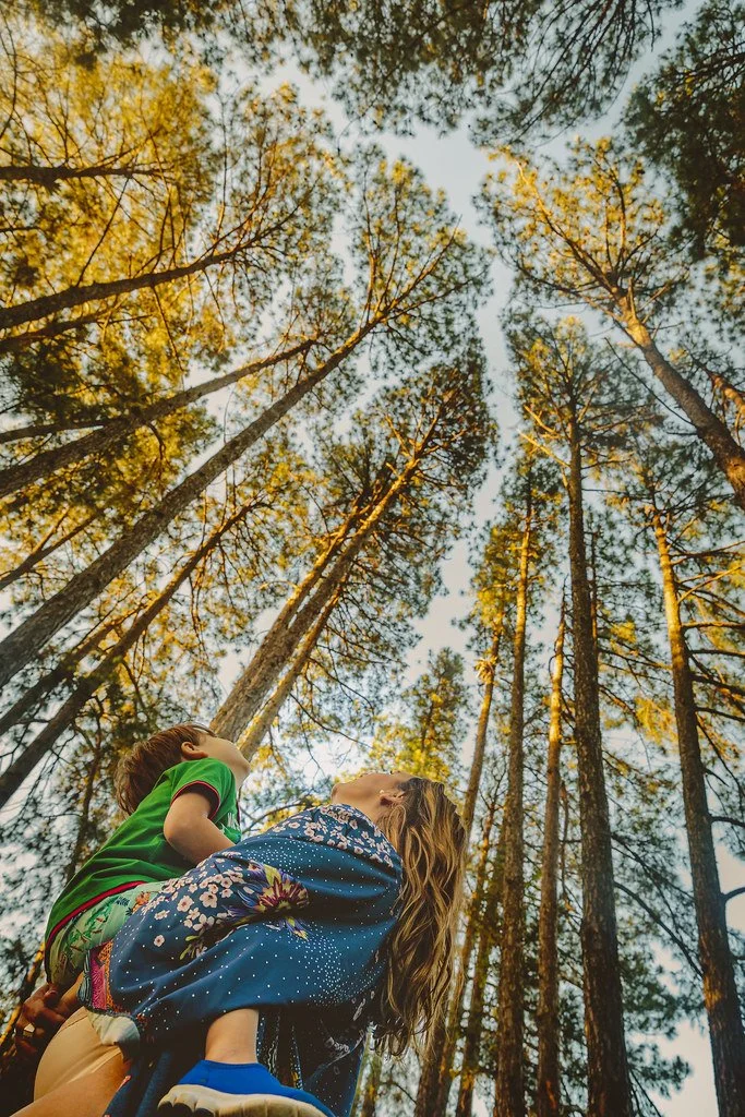 Crianças brincando em uma floresta, uma carregando a outra sob árvores altas com folhagem verde, durante o dia.