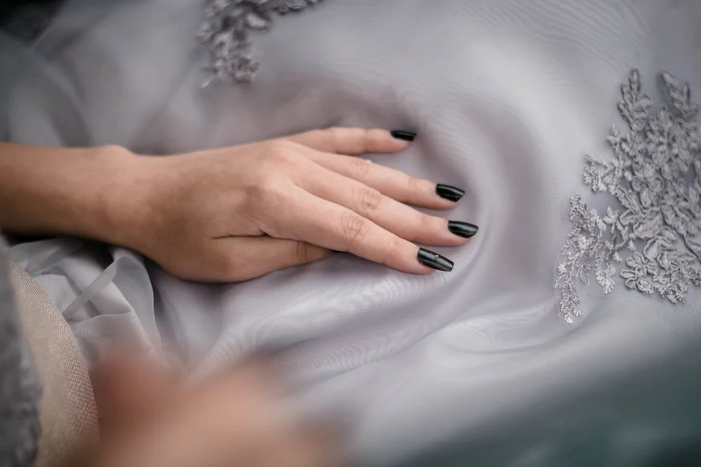 Mão feminina de unhas pintadas de preto repousando em tecido cinza com detalhes em renda.
