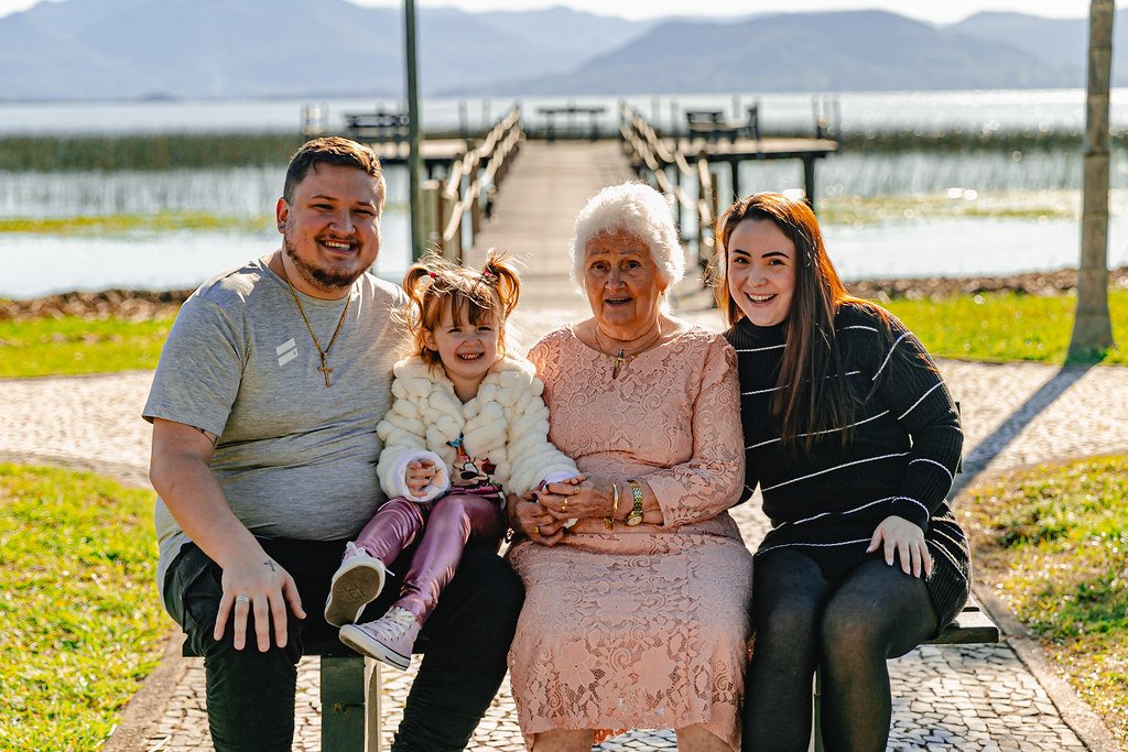 Grupo de cinco pessoas, incluindo uma criança, sentados em um banco ao ar livre com um pier ao fundo e uma paisagem de lago e montanhas.