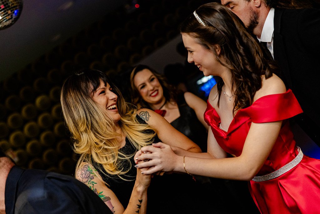Mulher sorrindo com cabelo loiro e tatuagens, usando vestido preto, interagindo com mulher de vestido vermelho, na companhia de outros amigos em ambiente interno.