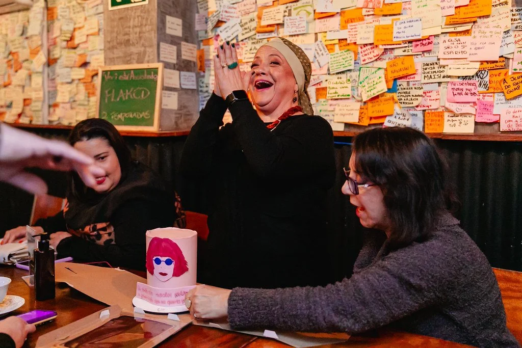 Mulher sorridente comemorando ao lado de duas outras mulheres em uma mesa, com parede cheia de bilhetes coloridos ao fundo.
