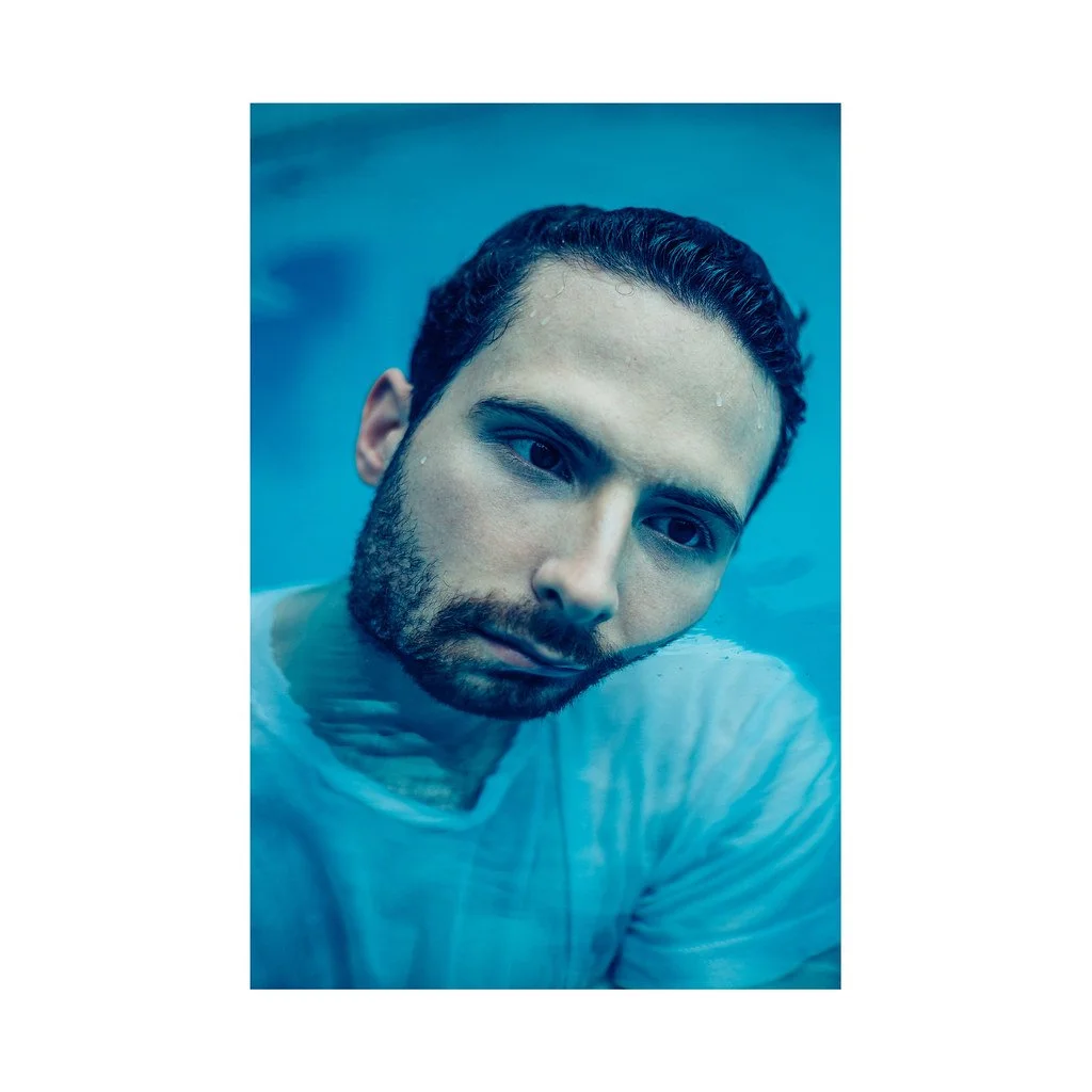 Retrato de um homem com barba, cabelo encaracolado, de roupa branca, com expressão séria, em ambiente com iluminação azul.