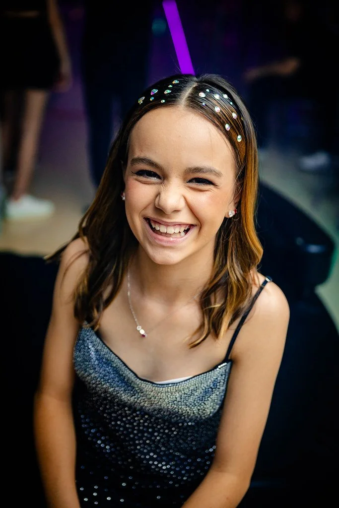 Menina sorrindo em ambiente de balada ou festa, usando vestido brilhante, brincos e colar, com decote de alça fina e cabelo solto.