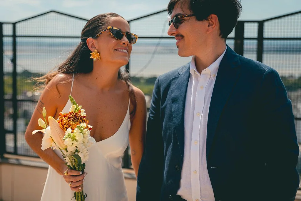 Casal sorridente em cerimônia ao ar livre, ela segurando buquê de flores, ambos de óculos escuros, vestindo roupas elegantes.