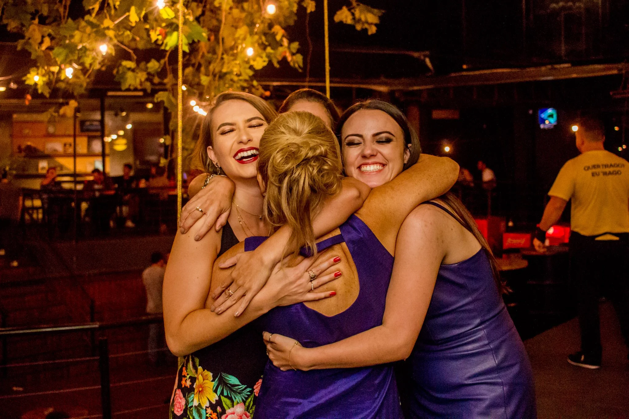 Grupo de quatro mulheres sorridentes abraçando-se, em ambiente de celebração ou festa, com iluminação quente e decorações ao fundo.