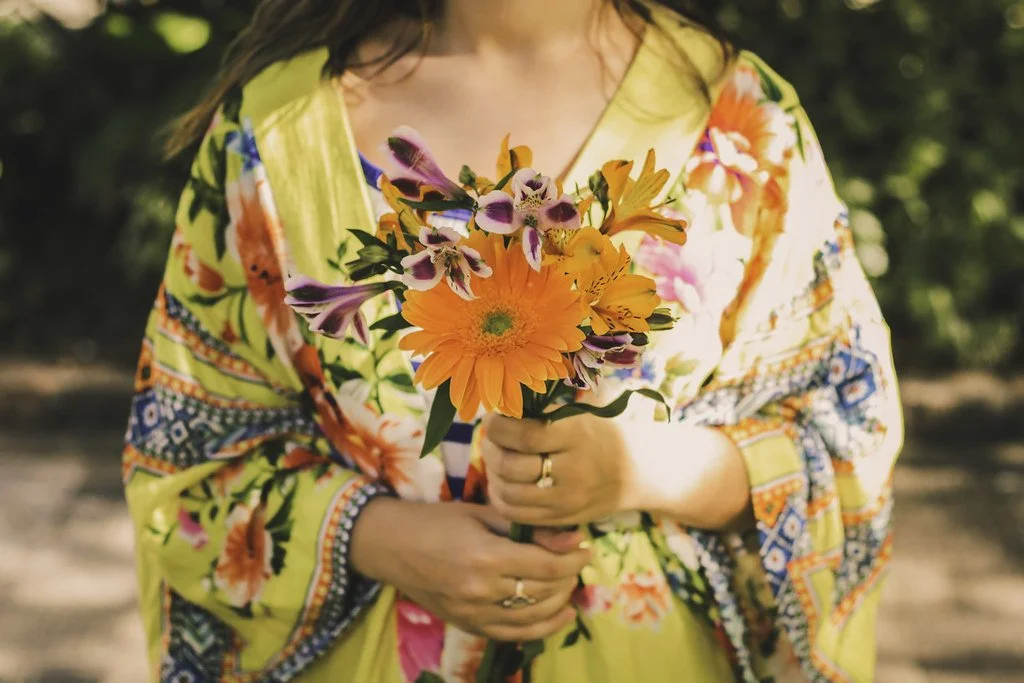 Pessoa segurando um buquê de flores coloridas, incluindo gérberas e orquídeas, enquanto veste uma roupa de estampas florais e vibrantes.