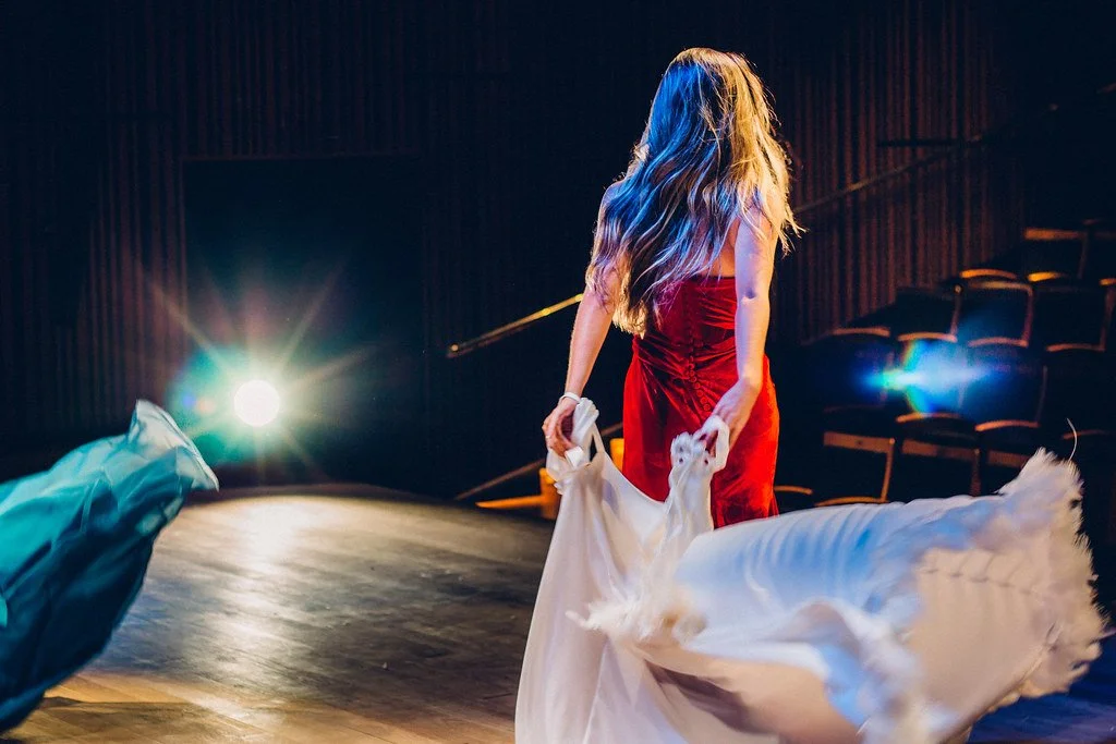 Mulher de vestido vermelho dançando na pista de teatro, com asas de anjo e iluminação teatral ao fundo.