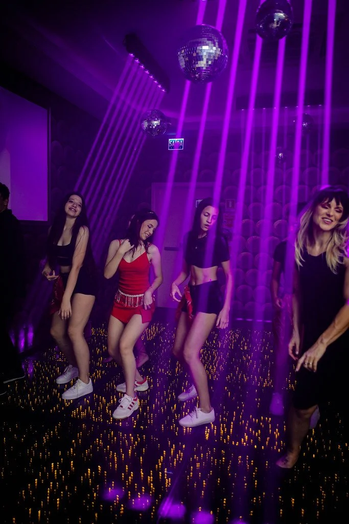 Grupo de jovens dançando em uma pista de dança com luzes coloridas, bolas de espelho e efeitos de luz roxa.