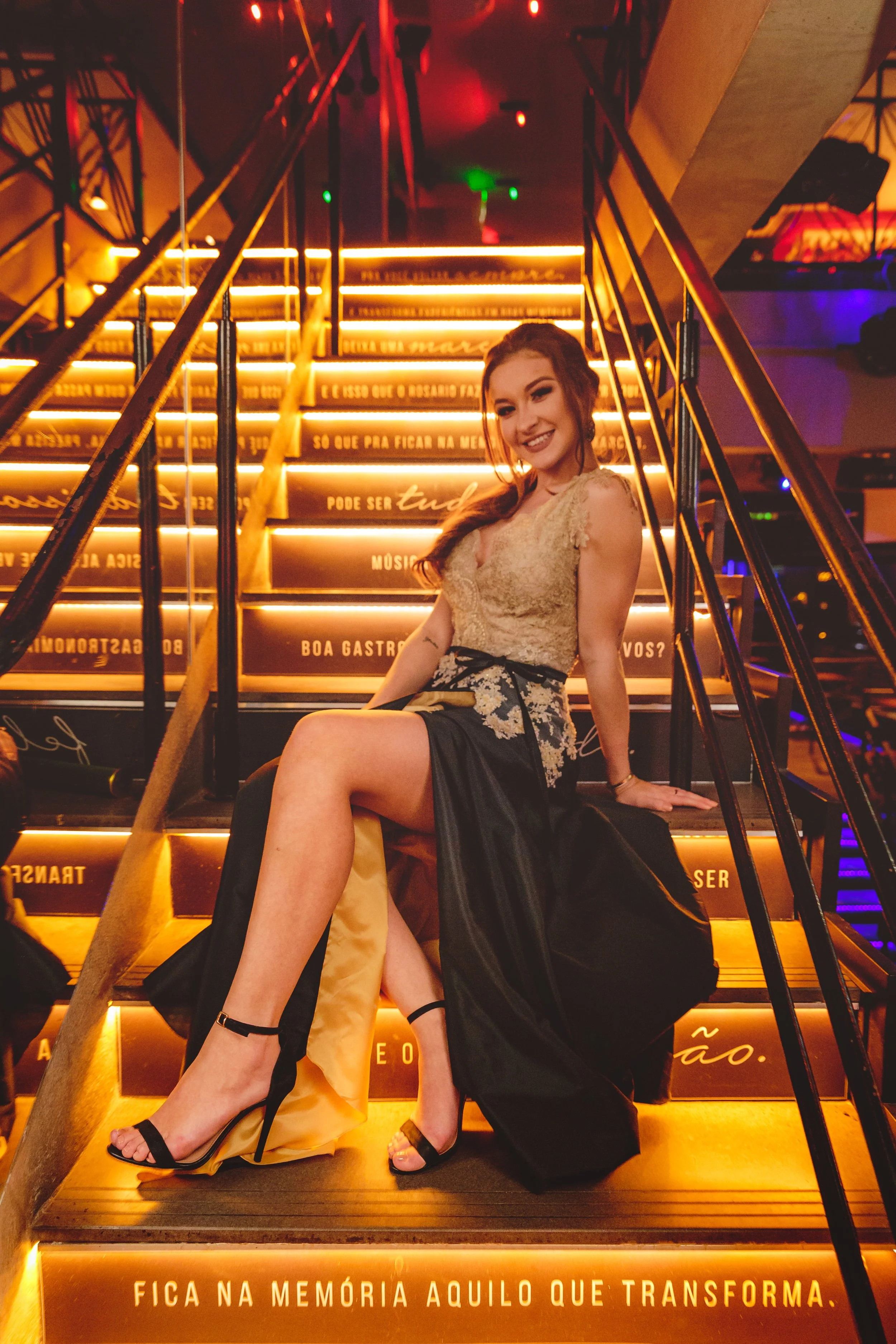 Mulher sorrindo sentada na escadaria iluminada com luzes de LED, de vestido elegante e salto alto, em ambiente sofisticado
