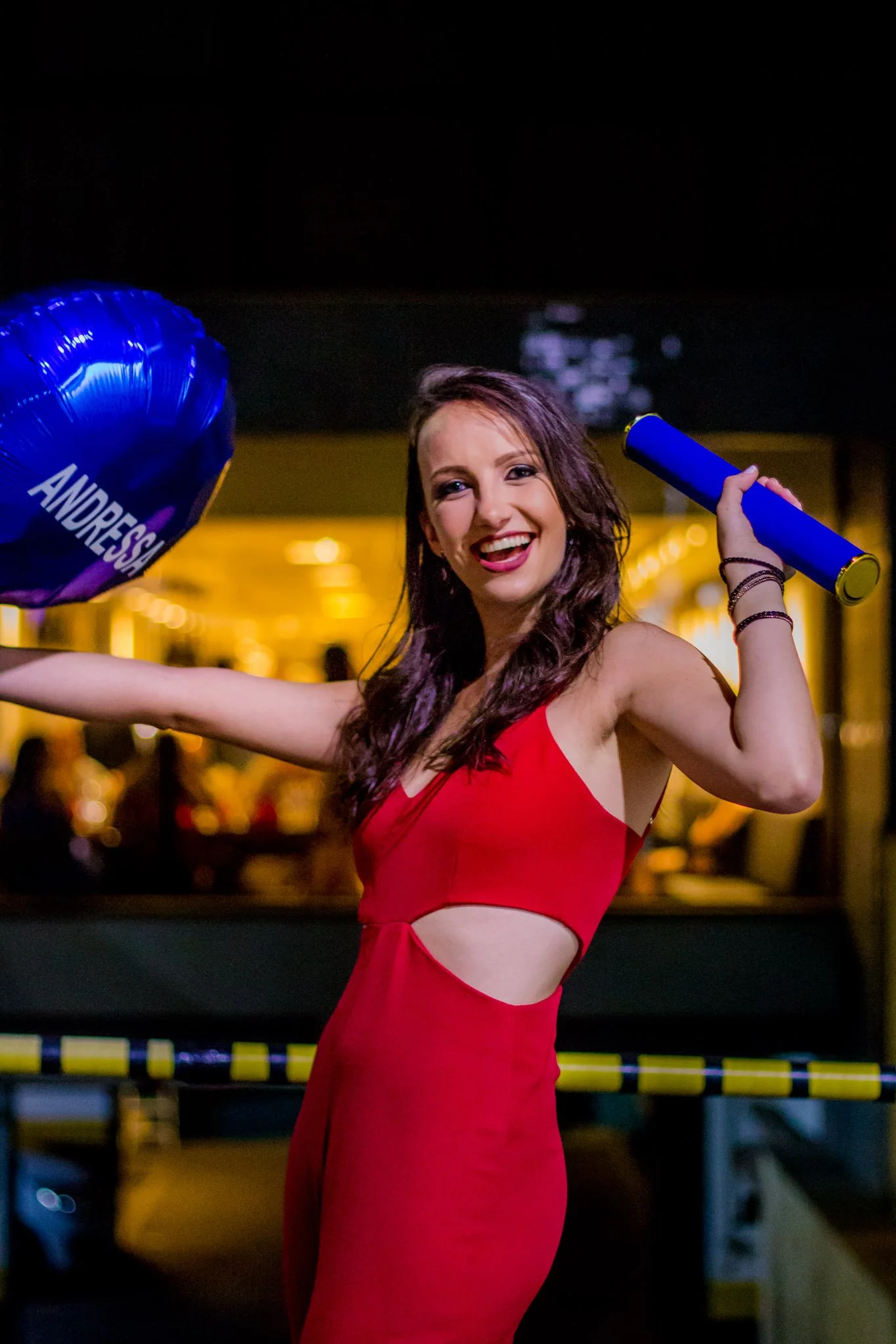 Jovem sorridente usando vestido vermelho, segurando um balão azul com o nome "Andressa" e um tubo azul com fundo de ambiente iluminado.