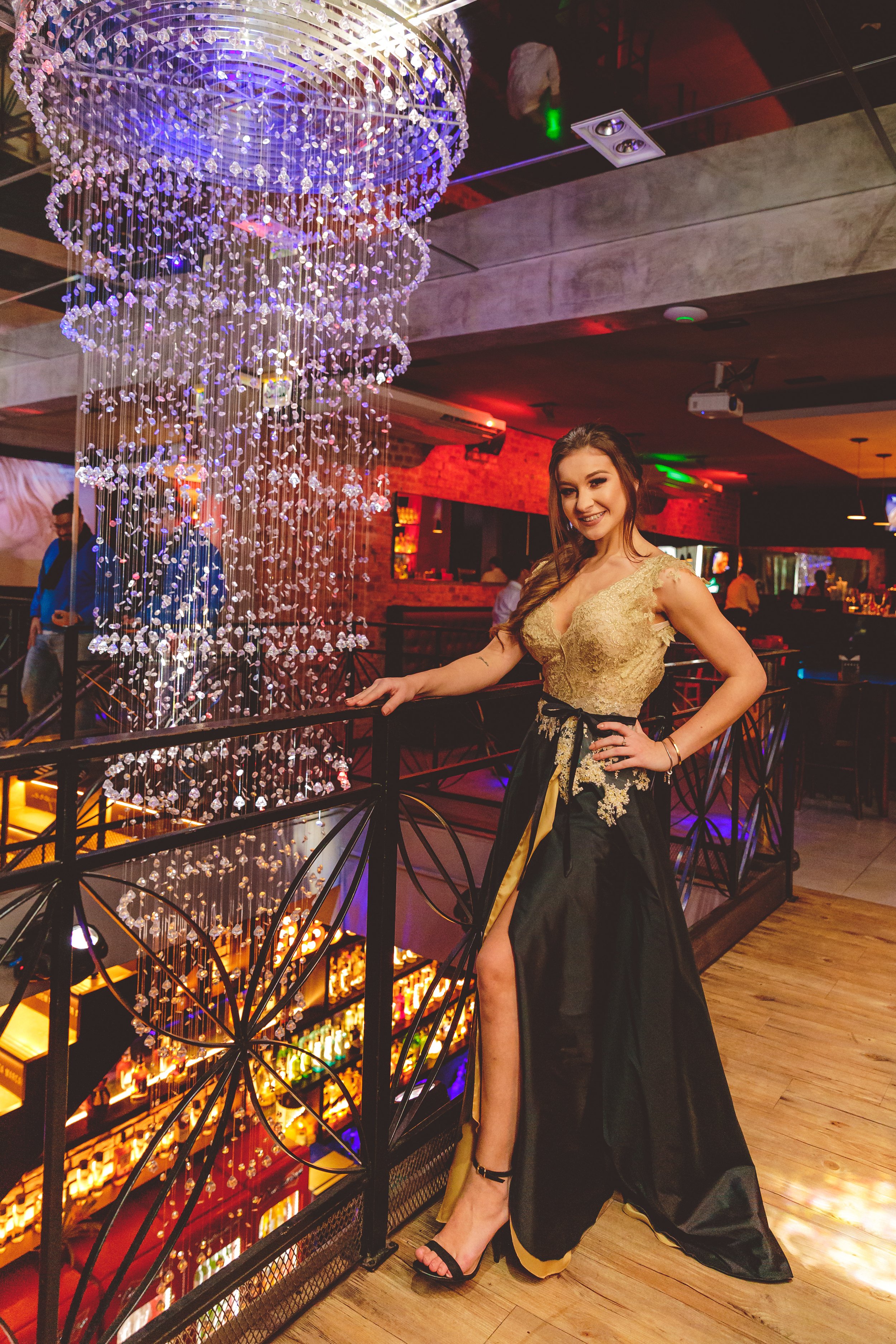 Mulher sorridente com vestido dourado e saia longa preta, posando ao lado de uma balaustrada decorada com um cristal pendurado, em um local com iluminação vibrante, possivelmente um restaurante ou bar.