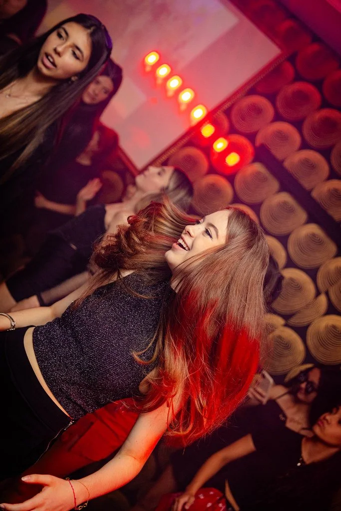 Homens e mulheres sorrindo e dançando em uma festa ou boate com iluminação vermelha e parede com padrão decorativo.