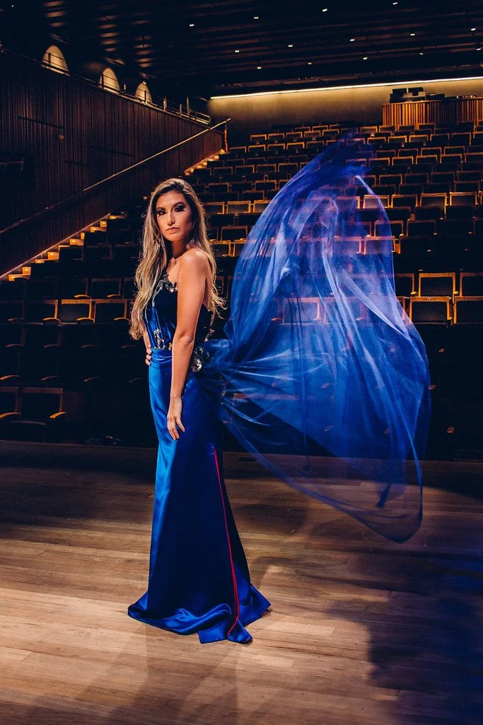 Mulher usando vestido azul na frente de uma plateia vazia em um teatro ou auditório, com cadeiras vazias ao fundo e iluminação suave.
