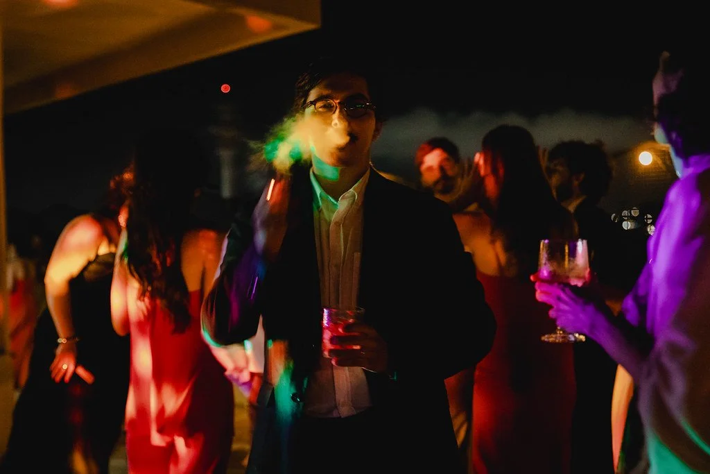Homem em traje formal e óculos preto fumando cigarro, ao fundo pessoas em uma festa à noite, algumas segurando taças, com iluminação colorida.