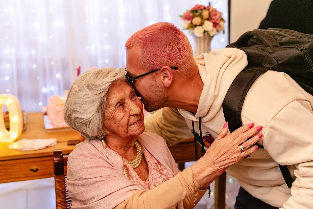 Homem idoso e jovem se beijando na testa em um ambiente com decoração de festa e uma mesa com flores ao fundo.