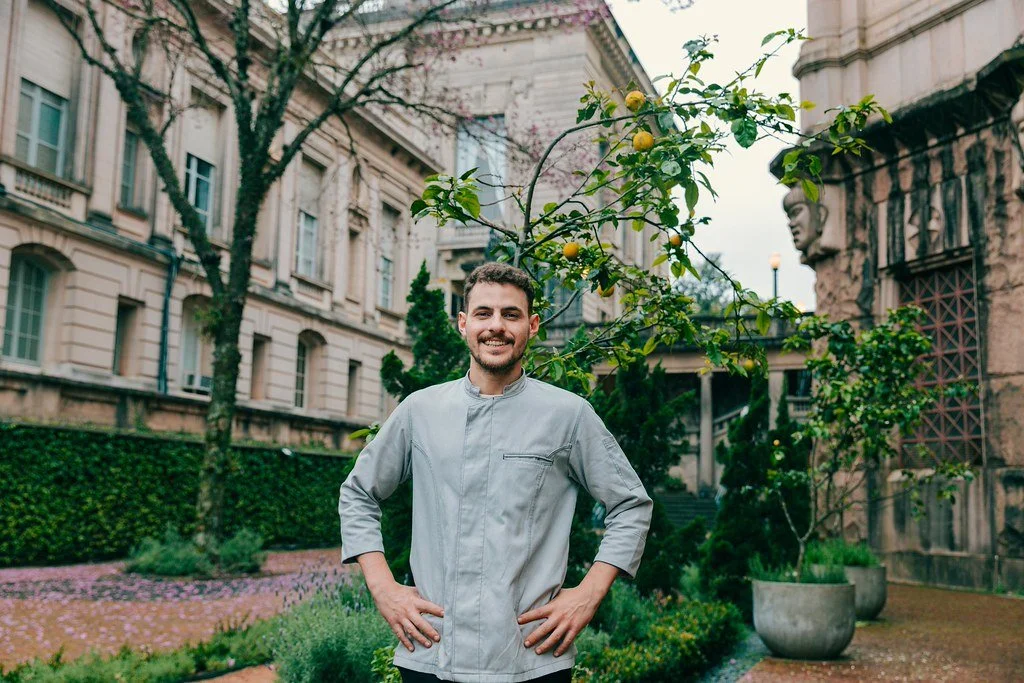 Homem sorridente de jaleco em jardim com árvores e plantas ao redor na cidade.