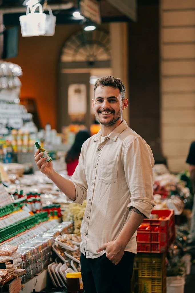 Homem sorridente segurando uma garrafa de molho em mercado, entre prateleiras de produtos alimentícios e utensílios de loja.