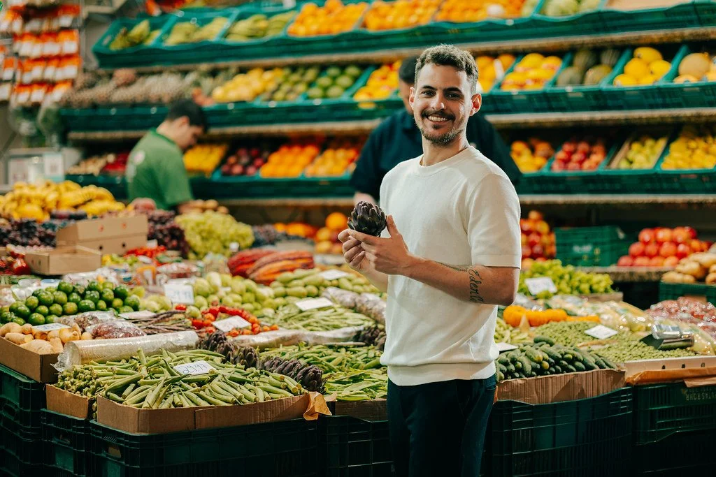 Homem sorridente segurando uma alcachofra em uma feira de hortifrúti com várias frutas e legumes ao fundo.