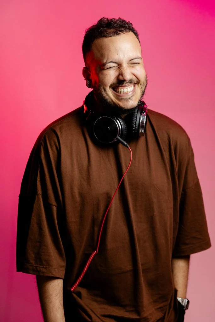 Homem sorrindo com fones de ouvido ao redor do pescoço, camisa marrom, fundo rosa.