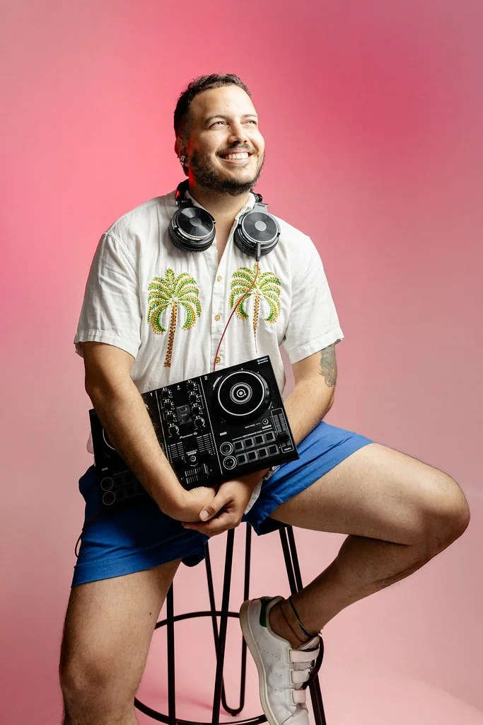 Homem sorridente, com barba e cabelo curto, usando uma camiseta branca com estampa de coqueiros, shorts azuis e tênis branco, sentado em um banco contra fundo rosa, segurando um aparelho de DJ, com fone de ouvido em volta do pescoço.