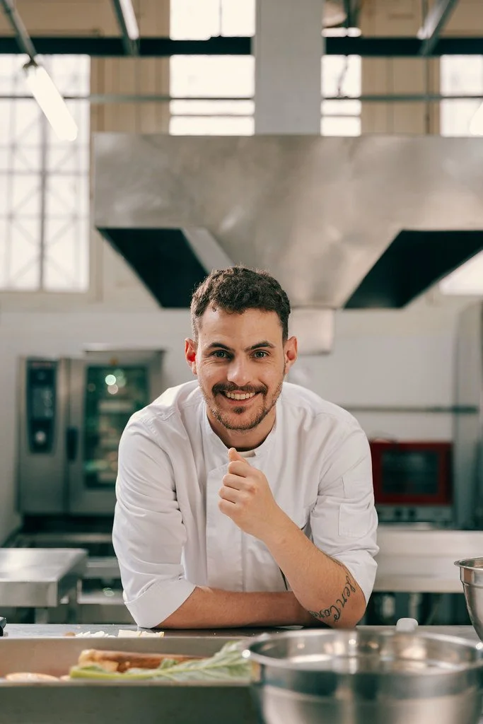 Homem sorridente em cozinha profissional, usando uniforme branco, com utensílios de cozinha ao redor.