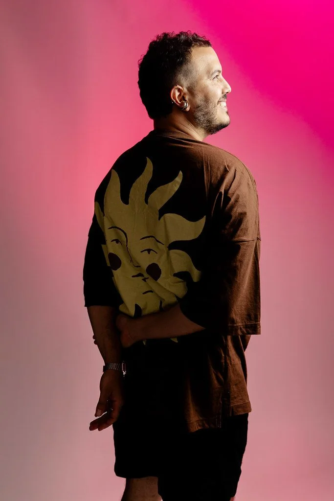 Homem de perfil sorridente usando camiseta marrom com estampa de rosto solar estilizado, fundo rosa