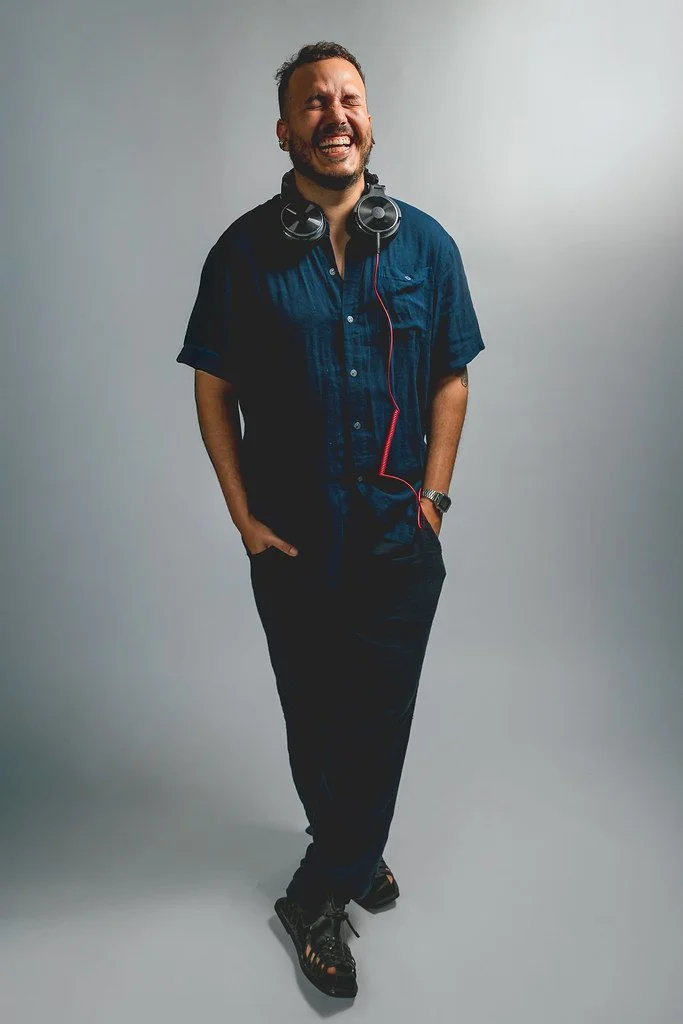 Homem sorrindo, usando camisa azul, calça preta, tênis e fones de ouvido ao redor do pescoço, posando com as mãos nos bolsos em frente a fundo cinza.