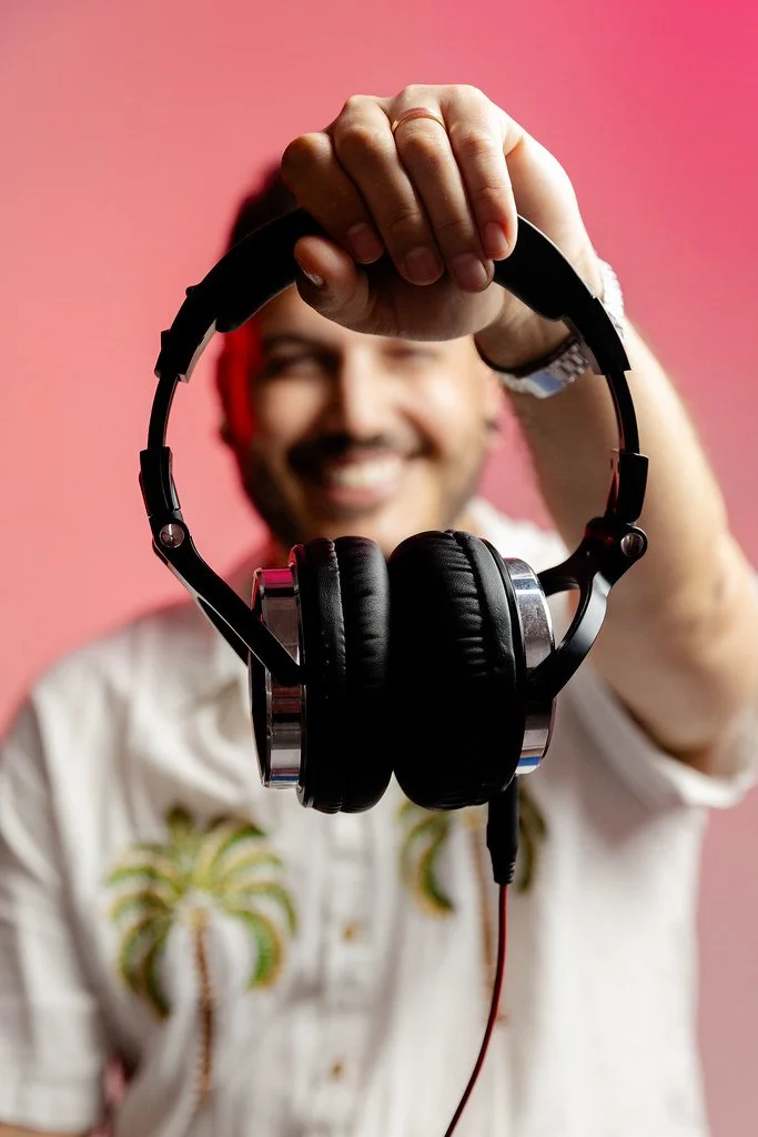 Homem sorridente segurando fones de ouvido em direção à câmera, com fundo rosa.