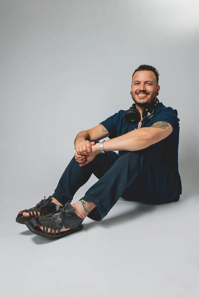 Homem sorridente sentado no chão com roupas casuais, acessórios de ouvido ao redor do pescoço, usando sapatos de sandália, com tattoos nos braços, em um fundo cinza.