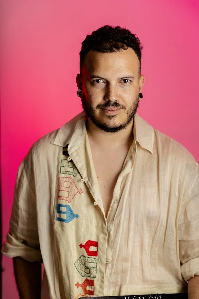 Homem com cabelo enrolado, barba, brinco na orelha e camiseta bege com desenhos coloridos, posando contra fundo rosa.