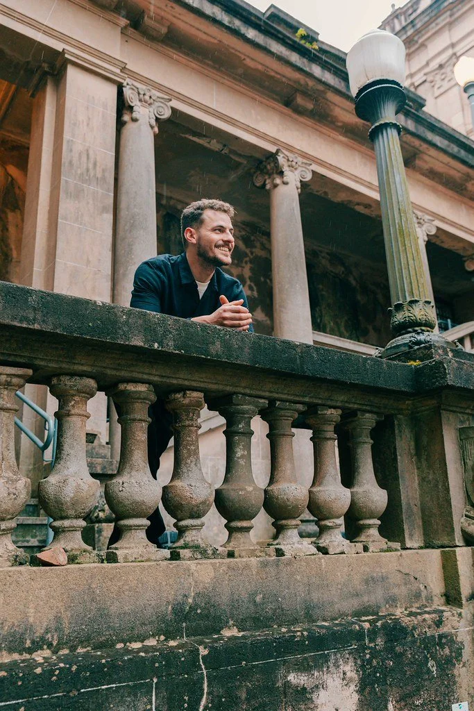 Homem sorridente apoiado num parapeito de uma varanda de um edifício antigo com colunas em estilo clássico, sob poste de luz de rua, durante um dia de chuva.