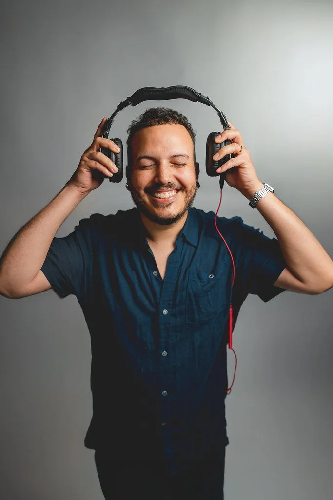 Homem sorridente usando fones de ouvido grandes, de cabelo curto e barba, segurando-os com as mãos, contra fundo cinza.