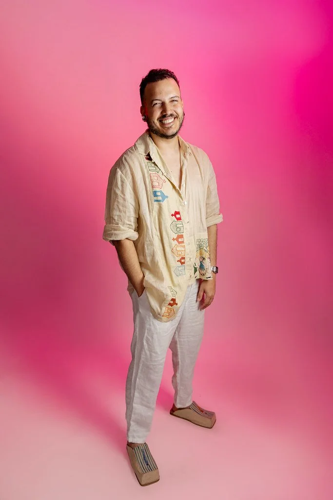Homem sorridente com cabelo curto e barba, vestindo camisa bege com bordados coloridos, calça branco e sandálias de salto alto com tiras azuis, posando contra fundo rosa.